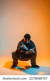 Handsome funny young man dancer in sunglasses and a hat in a down jacket with sneakers sits in a creative studio with orange and neon light - Shutterstock ID 2278088707