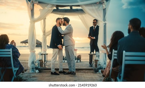 Handsome Gay Couple Exchange Rings And Kiss At A Beach Wedding Ceremony Venue At Sunset. Two Happy Men In Love Share Their Big Day With Diverse Multiethnic Friends. LGBTQ Relationship Goals.