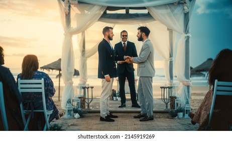 Handsome Gay Couple Exchange Rings And Kiss At A Beach Wedding Ceremony Venue At Sunset. Two Happy Men In Love Share Their Big Day With Diverse Multiethnic Friends. LGBTQ Relationship Goals.