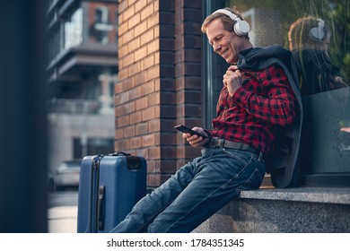 Handsome gentleman in headphones holding smartphone and smiling while enjoying favorite songs on the street