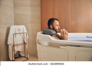 Handsome gentleman holding glass of alcoholic drink and smiling while relaxing in bathtub - Powered by Shutterstock