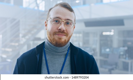 Handsome Ginger Creative Design Engineer With Beard And Glasses Poses For Camera Next To An Electric Car Chassis Prototype. In High Tech Laboratory Facility With Vehicle Frame.