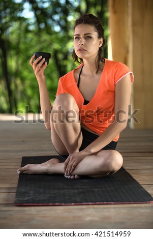 Handsome girl sits on the black yoga mat on the wooden terrace on the nature background. Her head is partially turned to the right, right foot stands on the mat, left leg lies on the mat and right