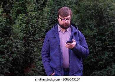 Handsome with glasses and a beard man using his phone, texting a message, sms on his mobile smartphone with smile standing on the street in winter near the trees.Concept: searching or social networks 