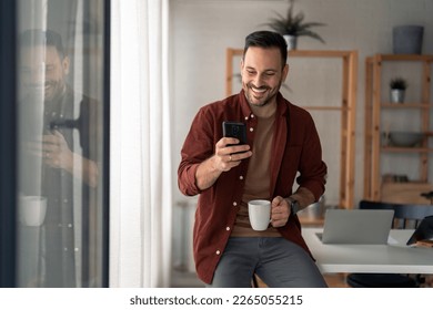 Handsome good-looking man, modern entrepreneur in stylish casual clothes cheerfully looking at mobile phone screen and holding white cup of coffee while sitting on the edge of the table in home office - Powered by Shutterstock