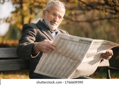 Handsome grandfather with a beautiful beard in a gray jacket sits on a bench in the park and reads a newspaper. Senior gray-haired man in glasses - Powered by Shutterstock