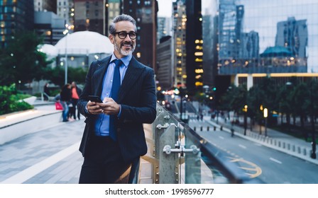 Handsome gray haired glad businessman with trendy haircut in black suit and in spectacles with smartphone by glass fence looking away on blurred background in New York