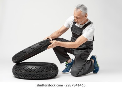 Handsome gray haired senior man choosing seasonal motorcycle tires, wearing overalls isolated on gray background. Repair, service concept - Powered by Shutterstock