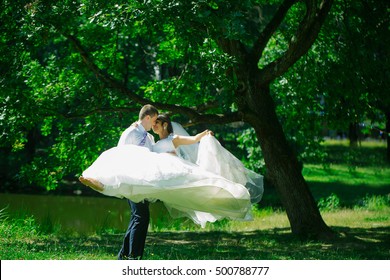 Handsome groom man circling with beautiful bride woman in wedding veil in hair and white flying dress on hands outdoors in summer park - Shutterstock ID 500788777