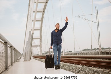 A handsome guy with a beard pulls a suitcase on the bridge next to the railway and waves