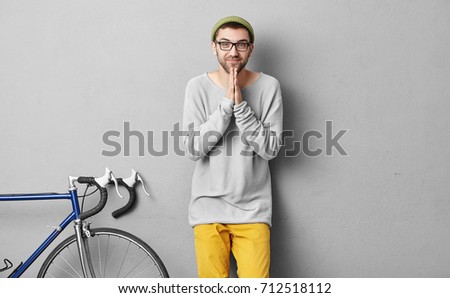 Handsome guy with beard, wearing hat, loose sweater and yellow trousers, keeping hands together having begging look, asking his friend to have ride on bicycles, isolated over grey background