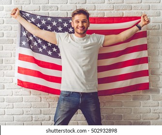 Handsome guy in casual clothes is holding American flag, looking at camera and smiling, on white brick wall background