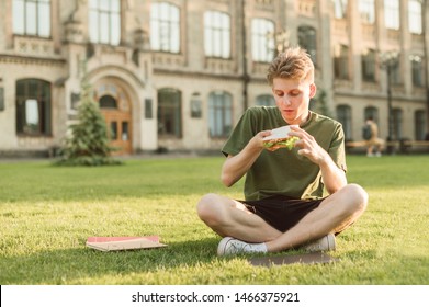 Handsome Guy In Casual Clothes Sitting On A Lawn On The Background Of A University Building With A Sandwich In His Hands And Going To Eat. Portrait Of A Hungry Student Eating A Sandwich At A Break.