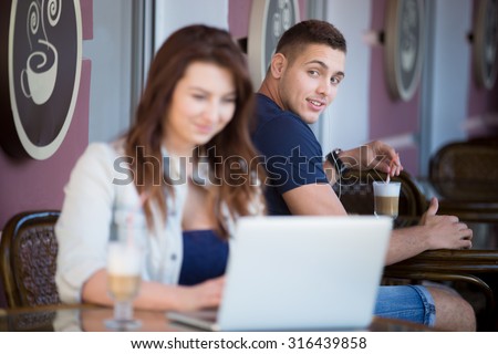 Handsome guy in casual clothes sitting in street cafe with a drink, looking with admiration at smiling beautiful young woman with laptop at the next table