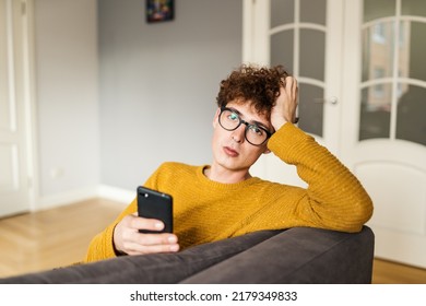 Handsome Guy In Eyeglasses And With Curly Red Hair Text Messaging On Cellphone And Looking At Camera. Man Holding Mobile Phone Sitting On Sofa At Home