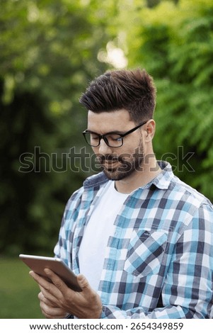 Handsome guy with glasess looking down at his digital tablet with interest while outdoors