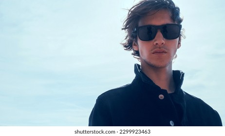 Handsome guy with long hair poses against sky with sun rays. Stylish man in black jacket looks at the camera, low angle shot. Young man with blown brown hair against the blue sky on summer sunny day. - Powered by Shutterstock