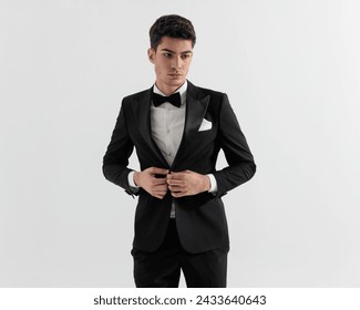 handsome guy model buttoning his black tuxedo while looking down to side on gray background