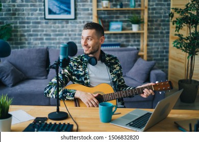 Handsome Guy Is Playing The Guitar Recording Audio Podcast In Studio Using Microphone And Laptop Sitting At Table Indoors Alone. People And Music Concept.