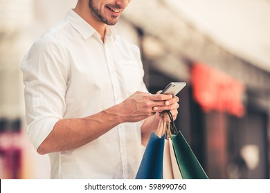 Handsome guy with shopping bags is using a mobile phone and smiling while doing shopping in the mall - Powered by Shutterstock