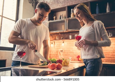 Handsome Guy Is Smiling And Cooking In The Kitchen While His Girlfriend Is Drinking Wine