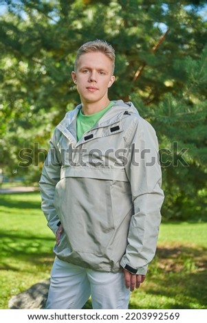 A handsome guy in stylish youth clothes stands in a green sunny park on the background of fir trees. Youth fashion. 