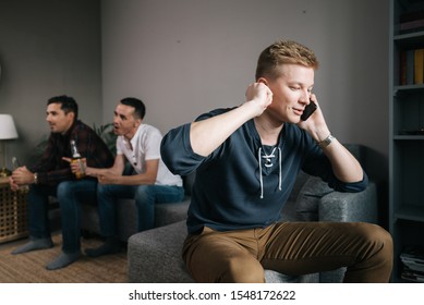 Handsome Guy Trying To Talk On The Phone. Behind Friends Are Watching TV. Group Of Friends Watching A Football Match On TV Against The Background Of Young Man Phone Conversation.