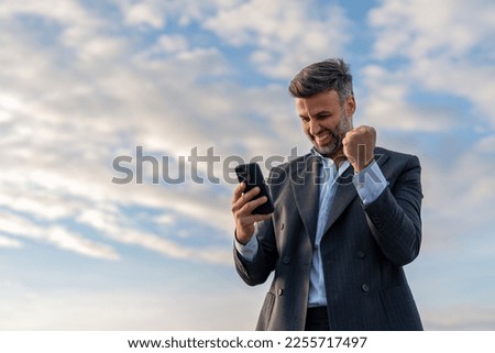 Handsome happy businessman feeling excited raising fist in yes gesture looking at cell phone celebrating work success, getting new approved job opportunity succeed in career and financial goals.