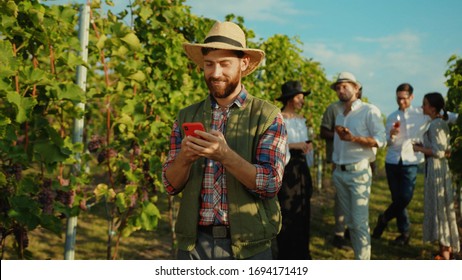 Handsome happy farmer in hat using application on smartphone in behind young luxury people tasting grapes and wine on vineyard. - Powered by Shutterstock
