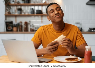 Handsome, happy Latin man holding tasty sandwich, eating, enjoying with closed eyes sitting in modern kitchen at home. Concept of food, freelancer working at home - Powered by Shutterstock