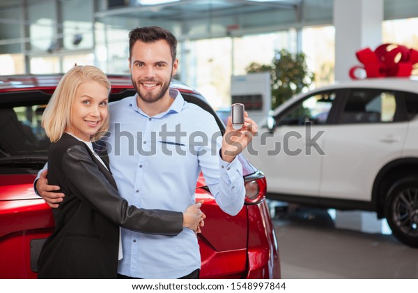 Handsome happy man hugging his lovely wife smiling\
to the camera with a car key in his hand, copy space. Couple\
celebrating buying new\
auto