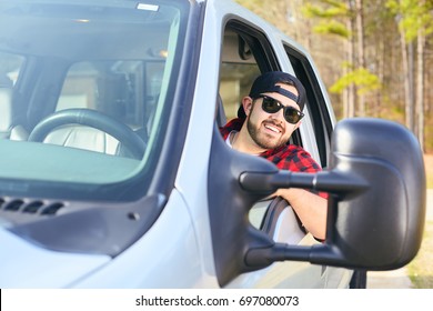 Handsome happy men driver with a beard smiling in the pickup car truck. Attractive male driving big vehicle, wearing hat, checkered shirt and black sun glasses. Sunny weather, summer