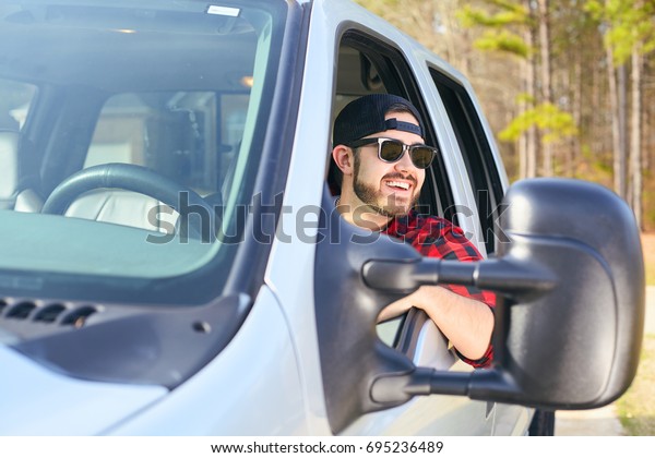 Handsome\
happy men driver with a beard smiling in the pickup car truck.\
Attractive male driving big vehicle, wearing hat, checkered shirt\
and black sun glasses. Sunny weather,\
summer