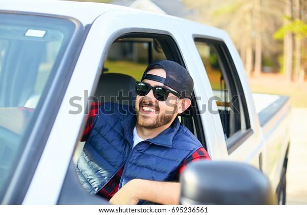 Handsome
happy men driver with a beard smiling in the pickup car truck.
Attractive male driving big vehicle, wearing hat, checkered shirt
and black sun glasses. Sunny weather,
summer