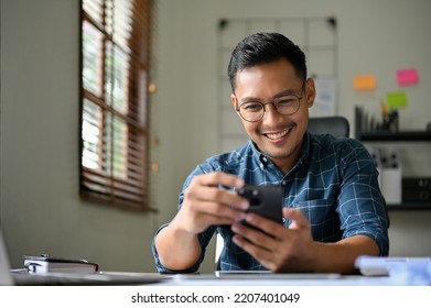 Handsome and happy millennial asian businessman or male office employee using his mobile phone at his desk. reading web blog, online news, sending email, message. - Powered by Shutterstock