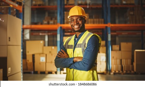 Handsome and Happy Professional Worker Wearing Safety Vest and Hard Hat Smiling with Crossed Arms on Camera. In the Background Big Warehouse with Shelves full of Delivery Goods. Medium Portrait