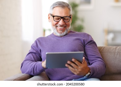 Handsome happy senior man using digital tablet while sitting on sofa at home, cheerful pensioner resting and relaxing in living room with electronic device, chatting with friends in social media - Powered by Shutterstock