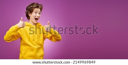 A handsome, happy teenager in trendy yellow clothes cheerfully smiles and shows his thumbs up. Purple background. Youth lifestyle and emotions. Copy space.