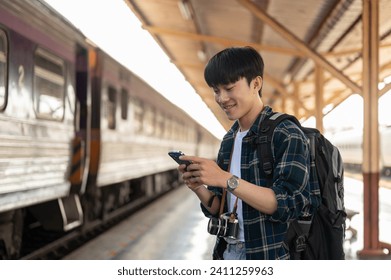 A handsome, happy young Asian man tourist backpacker using his smartphone, checking messages while standing at a platform in a railway station, waiting for his train. - Powered by Shutterstock