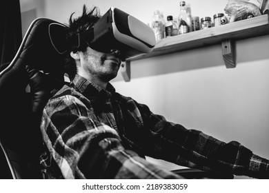 Handsome And Happy Young Latino Man With Plaid Shirt Playing With Virtual Reality Helmet (VR Glasses) On A Gamer Chair In Orange Bedroom On Home (in Black And White)