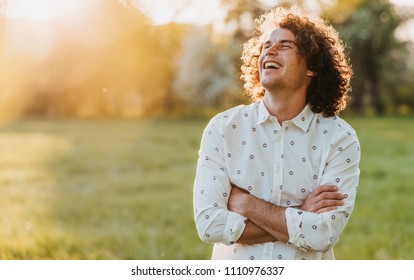 Handsome happy young male smiling with curly hair posing in the city park looking at the sky, cross his hands and dreaming. Copy space for your advertising. People, lifestyle and emotion concept
