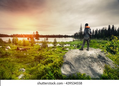 A handsome hiker is standing on the rock looking towards a view of beautiful nature in Grand Teton National Park.  This landscape has rocks, hiker, cloudy sky, and beautiful lake in distance.