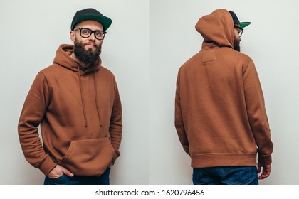 Handsome hipster guy with beard wearing brown blank hoodie or hoody from front and back and black cap with space for your logo or design on white background. Mockup for print
