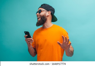 Handsome hipster guy with beard wearing orange t shirt and black cap closes eyes listens loud song in headphones, dances in rhythm of melody. Relax concept.