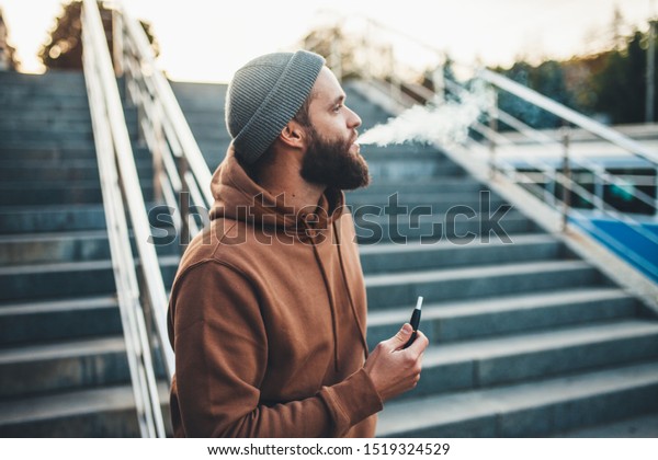 Handsome hipster guy with beard\
wearing brown blank hoodie smoking electronic\
cigarette
