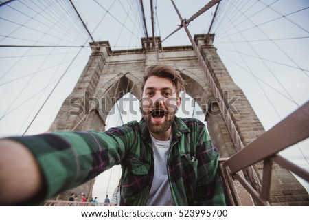 Handsome hipster guy taking selfie self-portrait on Brooklyn Bridge, New York. Cute bearded student takes funny picture for travel blog.