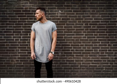 A handsome hipster guy wearing a blank gray t-shirt is looking aside while standing on a brick wall background. Empty place for the logo or design. Mock up.
