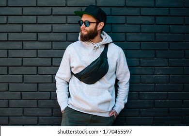 Handsome hipster guy wearing gray blank hoodie, black cap and black waist bag with space for your logo or design. Mockup for print