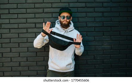 Handsome hipster guy wearing gray blank hoodie, black cap and black waist bag with space for your logo or design. Mockup for print