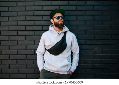 Handsome hipster guy wearing gray blank hoodie, black cap and black waist bag with space for your logo or design. Mockup for print